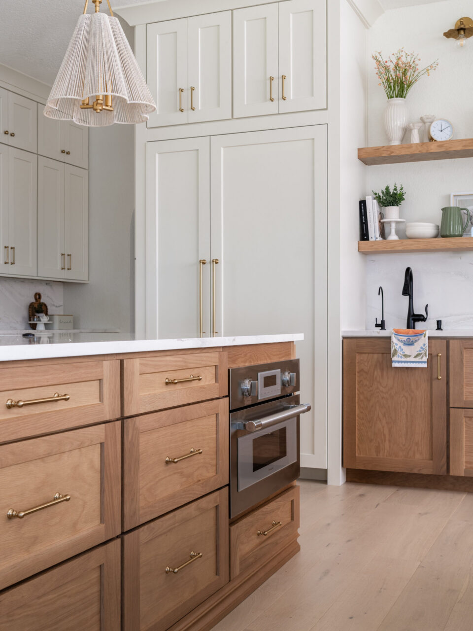 Modern kitchen with white upper cabinets and brass hardware above a light wood island and stainless oven beneath a marble countertop.