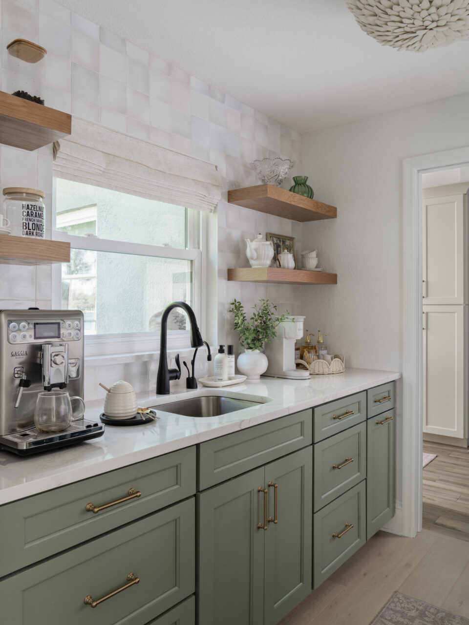 Modern kitchen with sage green cabinets, white marble countertop, and open wood shelves holding glassware and decor near a window.