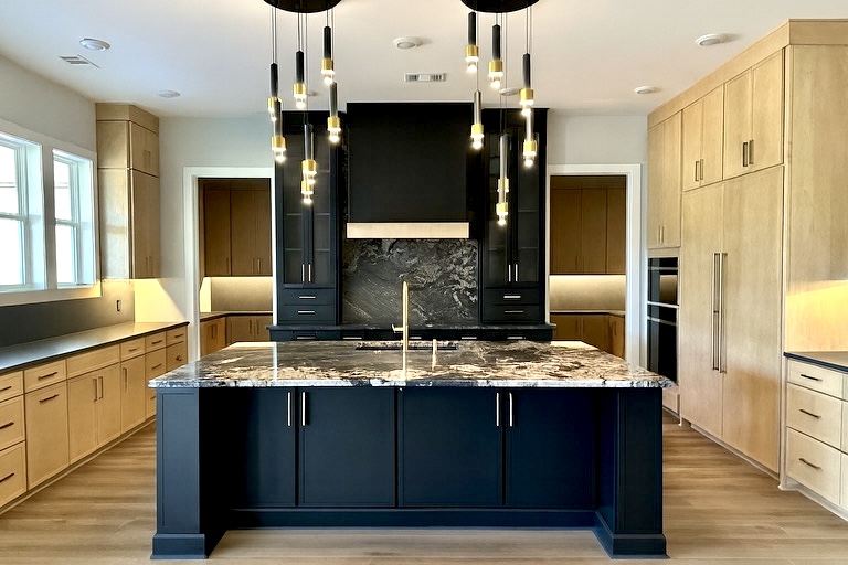 Modern kitchen with a dark blue island, marble countertop, and black range hood area framed by light wood cabinets.